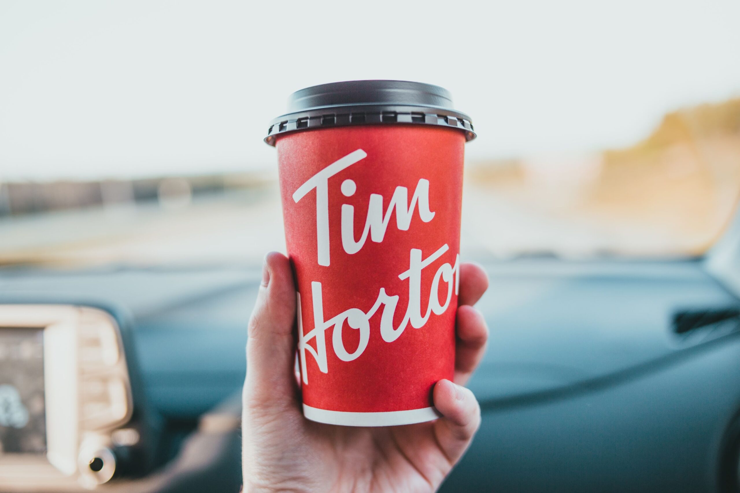 Photo of person holding a Tim Horton's coffee cup in front of a car dashboard. The Tim Horton’s Double Double is a hot coffee with two shots of cream and two packs of sugar and is now a huge part of Canadian culture!