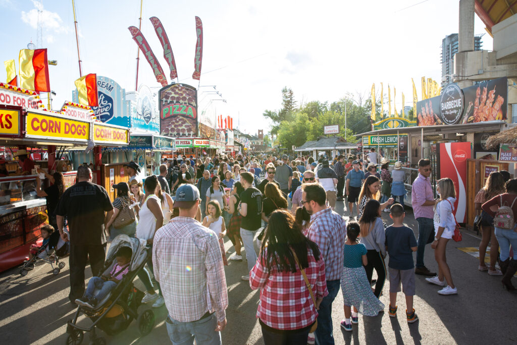 A group of people at the Calgary Stampede in front of food stands for pizza, corn dogs, barbecue, etc.
