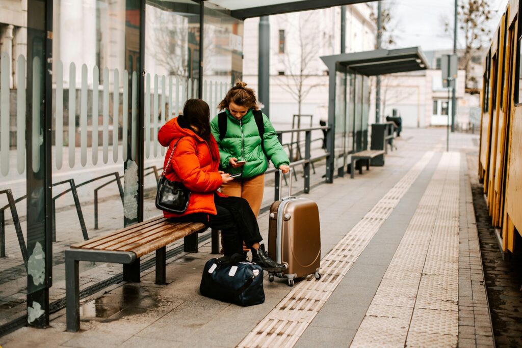 two women sitting on a bench with their luggage at a bus stop 