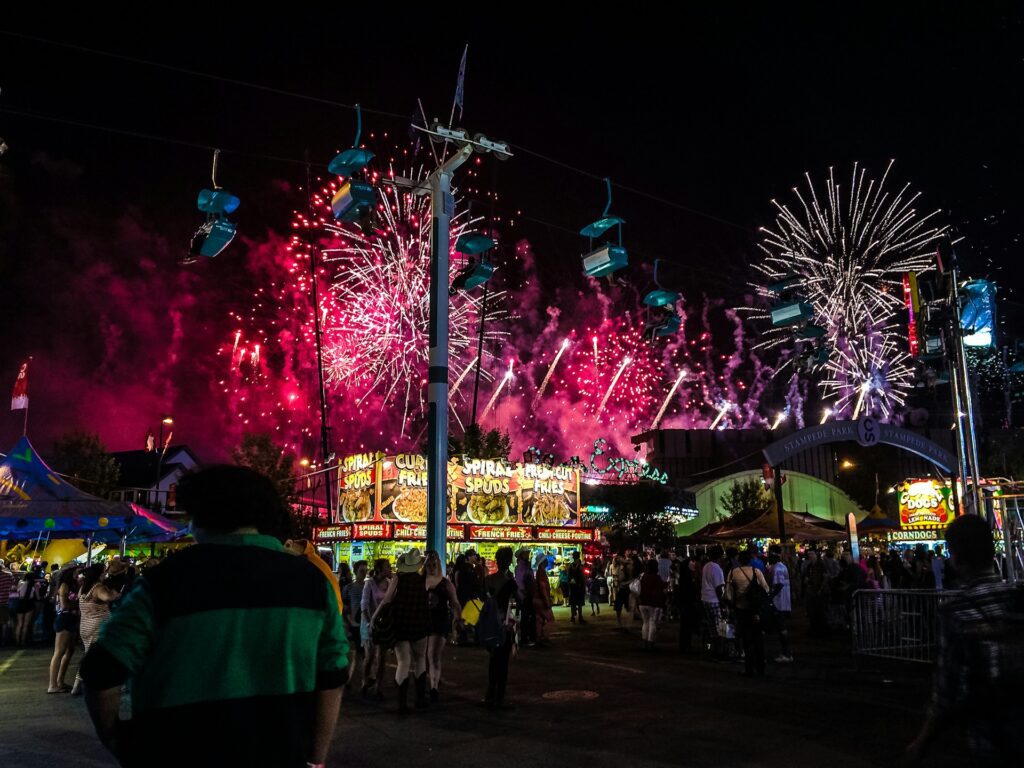 A crowd of people standing around a carnival at night watching fireworks  