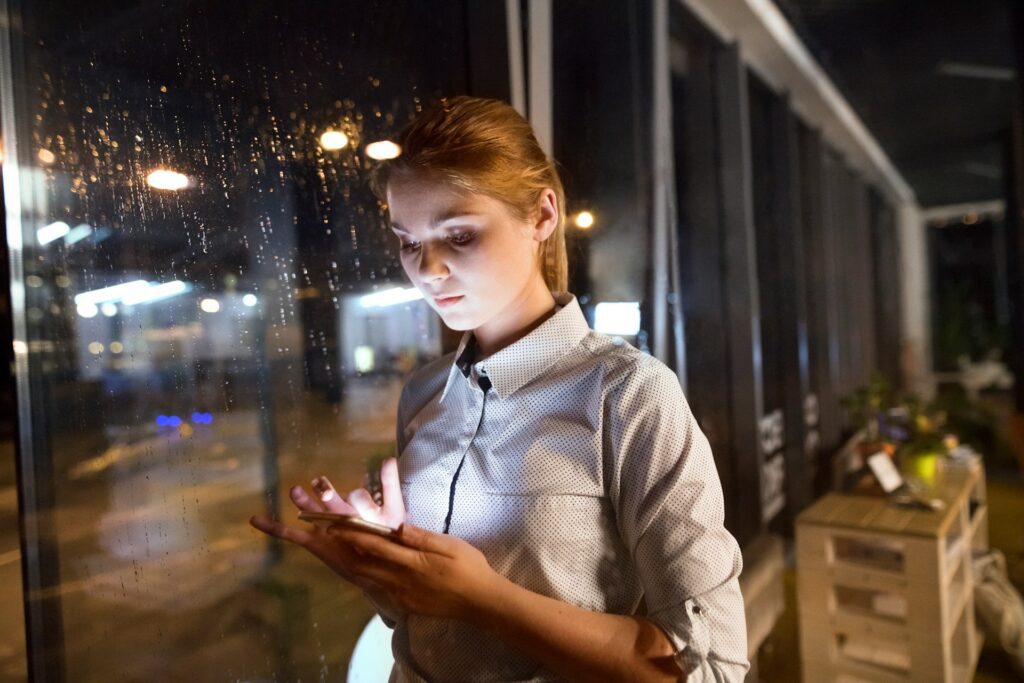 A woman checking her smart phone in the office next to a window which shows that it’s raining outside 